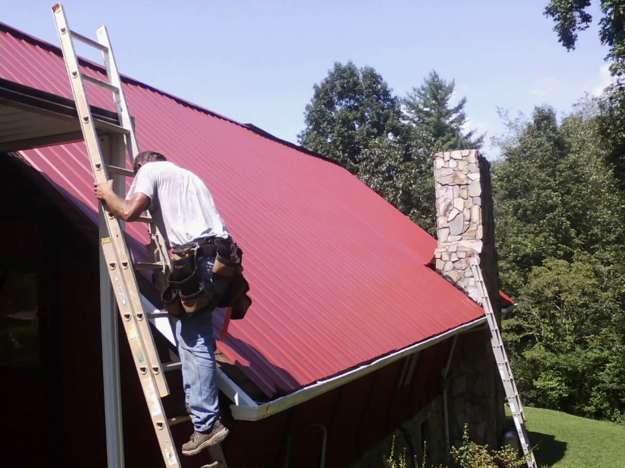 Roofer on a ladder beside a red metal roof and stone chimney — Soffit & Fascia Repair by Roofing Brothers in Plain Dealing